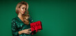 Elegant glamour blonde woman in evening sequin green dress is holding red Christmas gift on green background in studio.