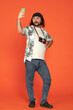A male traveler with a retro camera uses a smartphone to take a selfie or speak via video call. Full length man in the studio on an orange background. Vertical shot.