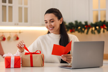 Sticker - Celebrating Christmas online with exchanged by mail presents. Smiling woman holding greeting card and opening gift during video call at home