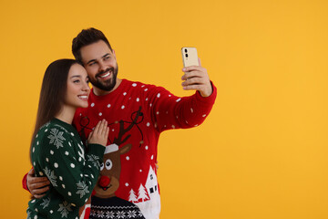 Wall Mural - Happy young couple in Christmas sweaters taking selfie on orange background. Space for text