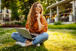 Curly-haired woman in casual clothes with a laptop sits on the lawn on a sunny day. Happy freelancer woman working outdoors and enjoying the weather.