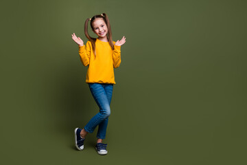 Sticker - Photo of cute cheerful schoolgirl with ponytails wear yellow pullover stand near empty space palms up isolated on khaki color background