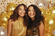 dos amigas de raza negra con vestidos dorados de lentejuelas con pello largo rizado castaño claro, sobre fondo con globos y confeti. concepto fin de año