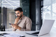 © Liubomir - Businessman thinking while sitting inside office at table, man is concentrating and seriously reading online using an application on phone, the young boss is holding a smartphone in his hands