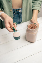 Wall Mural - Young woman in jeans and shirt holding measuring spoon with protein powder, glass jar of protein drink cocktail, milkshake or smoothie above white wooden table