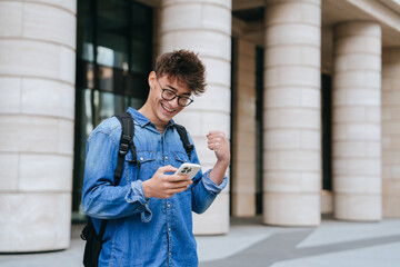 excited caucasian student guy in jeans shirt holds phone reads message clenches fist smiles wide. su