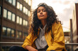 beautiful woman, teenager, low angle shot, in a big city, near a building, wearing a yellow jacket and hoodie, indian origin, sitting outdoors, smiling, pretty, diversity