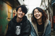 portrait of a couple outdoors, two asian teenagers laughing out loud, happy, white teeth, brown hair, in a street of a city, casual clothes, jackets with hood, green wall