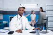 © DC Studio - Looking at the camera, African American male doctor sits at desk in hospital office. Portrait of black guy in a lab coat at clinic, with a patient in the background being aided by a nurse.