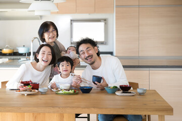 同居や二世帯住宅などのイメージ　おばあちゃんと子どもたちと孫の三世代家族のダイニングで食事のカメラ目線　右にコピースペースあり