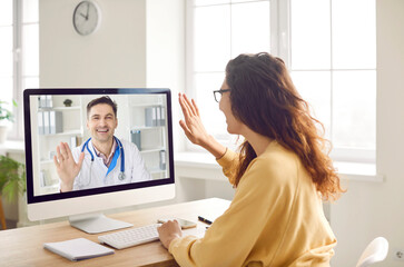 Patient says hello to online doctor when they start medical consultation via video call. Woman sitting in front of computer screen and waving her hand to greet her remote doctor. Telemedicine concept