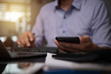 Businessman hand holding mobile smartphone on a table with a laptop at office...