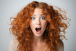 Emotional portrait of a woman, surprised or shocked Caucasian young curly redhead woman on a gray background looking at camera with big eyes