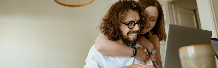 smiling man working on laptop while wife is hugging him at home. distance work concept