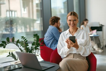 Wall Mural - Woman freelancer use mobile phone while working on laptop in modern coworking and looks camera