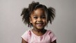 A radiant young black girl with curly pigtails and a bright smile wears a light pink shirt, her eyes sparkling with joy and innocence against a soft grey background
