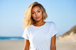 Woman with blonde hair standing on beach wearing white shirt.