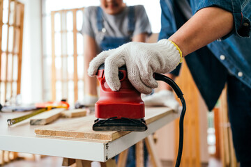 Close-up hand of an Asian father and son carpentry sanding timber plank to smooth with an electric sander machine together carefully at the home studio. Carpentry working at a home workshop studio.