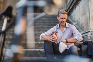 Wall Mural - Man sitting on stairs with takeaway coffee in the city checking the time