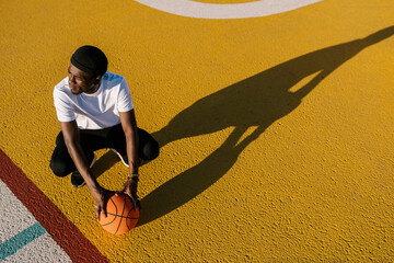 Canvas Print - Thoughtful young man holding basketball crouching on sports court during sunny day