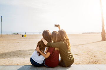 Wall Mural - Rear view of three female friends sitting at the beach taking a selfie