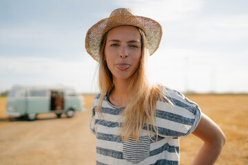 Poster - Portrait of young woman at camper van in rural landscape sticking out her tongue