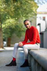 Poster - Portrait of fshionable young man wearing red pullover and sunglasses sitting on stone bench