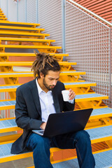 Wall Mural - Young businessman with dreadlocks sitting on stairs using laptop