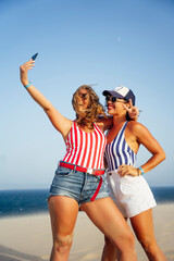 Wall Mural - Cheerful female friends taking selfie while standing at beach against clear sky