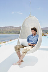 Man sitting in hanging chair above swimming pool using laptop