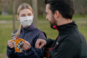 Wall Mural - Young woman giving face mask to man while sitting at park during quarantine