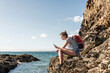 Young woman sitting on a rocky beach, using smartphone