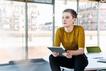 Thoughtful businesswoman sitting on desk in office holding tablet