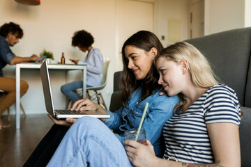 Wall Mural - Two smiling young women sitting on floor sharing laptop with friends in background