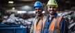 Indian worker in protective gear standing by waste paper in recycling station, with smiling colleague beside them.