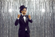 Happy elegant young ethnic man in party glasses, stylish classic black suit, white shirt, bowtie and top hat standing on shiny studio background, smiling and pointing index fingers at camera