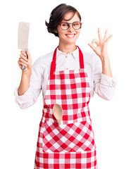 Sticker - Beautiful young woman with short hair wearing professional baker apron holding knife doing ok sign with fingers, smiling friendly gesturing excellent symbol