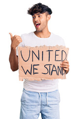 Poster - Young hispanic man holding united we stand banner pointing thumb up to the side smiling happy with open mouth