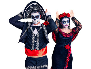 Wall Mural - Young couple wearing mexican day of the dead costume over background doing bunny ears gesture with hands palms looking cynical and skeptical. easter rabbit concept.