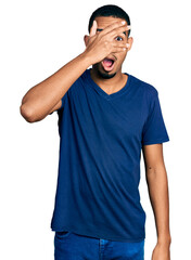 Wall Mural - Young african american man wearing casual t shirt peeking in shock covering face and eyes with hand, looking through fingers with embarrassed expression.