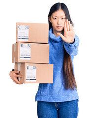 Poster - Young beautiful chinese woman holding delivery cardboard boxes with open hand doing stop sign with serious and confident expression, defense gesture