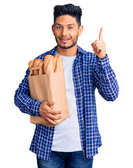 Wall Mural - Handsome latin american young man holding paper bag with bread pointing finger up with successful idea. exited and happy. number one.