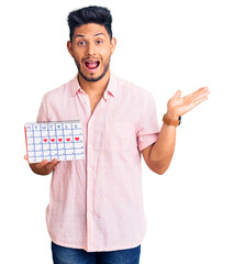 Sticker - Handsome latin american young man holding heart calendar celebrating victory with happy smile and winner expression with raised hands