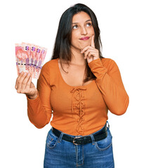 Sticker - Beautiful hispanic woman holding south african 50 rand banknotes serious face thinking about question with hand on chin, thoughtful about confusing idea