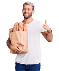 Canvas Print - Young handsome blond man holding paper bag with bread smiling happy and positive, thumb up doing excellent and approval sign