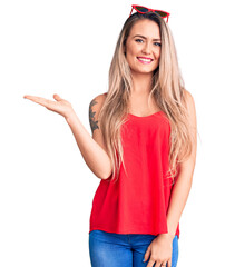 Wall Mural - Young beautiful blonde woman wearing sleeveless t-shirt and sunglasses smiling cheerful presenting and pointing with palm of hand looking at the camera.