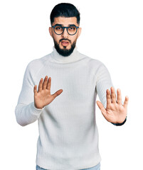 Wall Mural - Young arab man with beard wearing elegant turtleneck sweater and glasses moving away hands palms showing refusal and denial with afraid and disgusting expression. stop and forbidden.