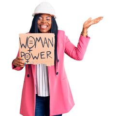 Wall Mural - Young african american woman wearing architect helmet holding woman power banner celebrating victory with happy smile and winner expression with raised hands