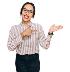 Wall Mural - Young hispanic girl wearing casual clothes and glasses amazed and smiling to the camera while presenting with hand and pointing with finger.