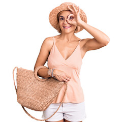 Canvas Print - Young blonde woman wearing summer hat holding bag smiling happy doing ok sign with hand on eye looking through fingers